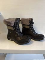 Side angled photo of both UGG Adirondack II women's boots showing black leather and synthetic body, fur collar, and treaded rubber sole.