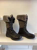 Side view of the tall UGG Adirondack boots showing black leather lower portion, brown suede upper, shearling cuff, and lace-up front.