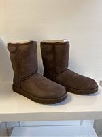 Side and angled view of pair of brown UGG Classic Short ladies boots size 11, showing outer suede material and plush lining at the top.