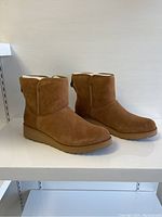 Side view of the pair of new UGG light brown suede boots on a white shelf