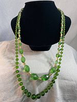 Two vintage green glass bead necklaces displayed on black necklace bust showing faceted and rounded beads.