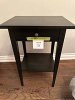 Front view of one black wooden bedside table showing drawer and bottom shelf with handwritten dimensions taped to drawer front.