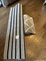 Two rolled area rugs. Larger rug has black and gray stripes, smaller rug has beige and gray geometric pattern.