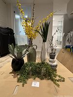 Six assorted vases with various artificial plants and greenery arranged on a wooden surface.