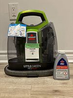 Front view of Bissell Little Green ProHeat cleaner with attached hose and cleaning formula bottle beside it on the floor, showing brand and product labels.
