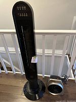 Full view of black NOMA oscillating tower fan standing next to silver and black Vornado whole room heater on hardwood floor.