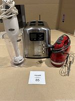 Three small kitchen appliances displayed: Oster immersion blender with clear measuring cup, Cuisinart brushed stainless steel 2-slice toaster, and red KitchenAid hand mixer with beaters.