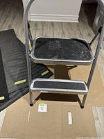 Photo showing metal two-step step stool with black textured steps, some pet fur on top surface, standing on cardboard on laminate floor.