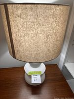 Front view of the lamp showing light brown fabric drum lampshade and white ceramic base with geometric design.