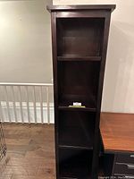 Front view of a tall, dark wooden bookshelf with three shelves, placed next to a wooden desk on a hardwood floor.