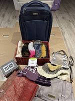 Overview of lot showing various scarves folded in a basket, hats, a basket with rolled scarves, fabric pieces in warm colors, a beige and silver purse, and a black Samsonite suitcase in the background.