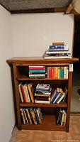 Front view of solid wood 3-shelf bookcase filled with assorted books against a white wall.