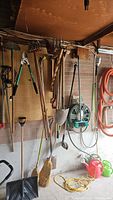 Garden tools hanging on wall including shovels, rakes, pitch fork, broom, and garden hose reel.