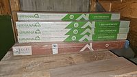 Seven unopened boxes stacked: six Nirvana Plus laminate flooring boxes in green and white, and one brown St. James Collection V-Groove box