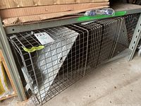 Side view of the metal mesh live animal trap positioned on a garage floor beneath a metal shelf, showing the wire grid and spring-door mechanism.