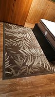 Full view of the rectangular outdoor rug placed beside a bed and wood-paneled wall and floor. Shows leaf motif design and color scheme.