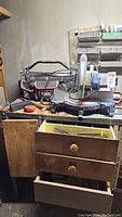 Ryobi TS230 table saw sitting on a wooden surface with Husky tool bag behind and open wooden drawers below containing more tools and parts.