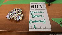 Top-down image of vintage Sherman rhinestone brooch sitting on wooden surface with MaxSold lot tag.
