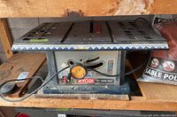 Front view of Ryobi 10 inch table saw on shelf showing top surface, cord, and brand label.