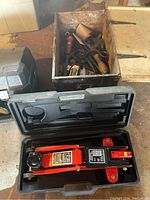Photo showing red hydraulic trolley jack in black hard plastic case alongside old wooden box filled with various hand tools.