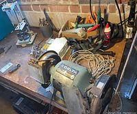 Side and angled view of Delta 6 inch bench grinder on workbench showing grinding wheels, power cord, and surrounding tools like pliers and mallet.