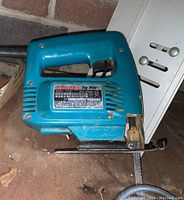 Makita jig saw in teal blue with black blade on wooden surface near white cabinet.