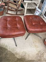 Pair of square swivel ottomans with brown leather tufted cushions and metal legs, positioned outdoors on concrete floor among other furniture items.