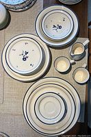 Top-down photo showing a stack of plates, two bowls, two mugs, and a small pitcher all featuring blue floral patterns on cream-colored ceramic.