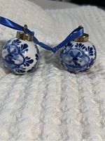 Two small blue and white Delft-style glazed ceramic ball ornaments with blue ribbons for hanging, featuring classic windmill and floral designs