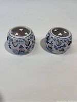 Pair of round white ceramic tea light holders with hand painted blue floral and heart patterns. Both have heart shaped cut outs at the top for ventilation.