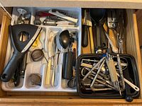 Top-down view of an open drawer divided into compartments containing assorted kitchen tools including black plastic vegetable peeler, metal spoon, corkscrews, can opener, and utensils with black and white handles.
