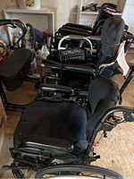 Photo showing four black manual wheelchairs lined up with their armrests and footrests visible, along with two walkers in the background.