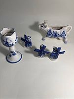 Photo showing five Delft blue ceramic items: cow creamer, goblet, three small windmill figurines. White ceramic with blue traditional designs, arranged on neutral background.