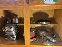 Mixed pots and pans arranged on two shelves in wooden cabinet, showing variety of cookware items stacked and nested.