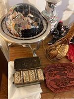 Table displaying a black globe on a metal stand, decorative ceramic vase, woven rooster figurine, red decorative tray, collectible dolls in traditional dresses, and small ceramic figurines arranged closely together.