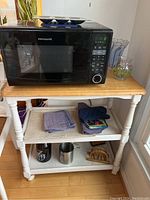 White rolling kitchen cart with wooden top and black Frigidaire microwave on top, folded kitchen towels on middle shelf, and items on lower shelf visible.