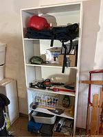White shelf unit filled with various tools and accessories including gas can, black bag, boxes, hand tools, and basket.