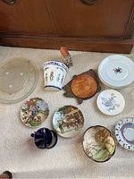 Photo showing various china serveware items including platter, plates with painted scenes, ceramic pitcher, wooden trivet, small cup, and house-shaped figurine.