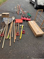 All garden and hand tools including rakes, shovels, red snow shovel, axes, sledgehammer, garden auger, aerobic stepper box, and shoe rack shown outdoors on paved surface.