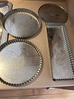 Three vintage metal baking pans on a counter, two circular with fluted edges and one rectangular with fluted edges, all showing use and needing cleaning