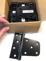 Box of 10 black Norton gate hinges showing stacked hinges inside the box and one hinge outside the box with a hand holding the box.