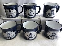 Photo of six navy blue ceramic mugs arranged on their boxes, three on top and three at bottom, showing three designs: lighthouse, anchor, sailor.