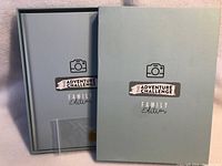 Closed light blue hardcover photo album titled 'The Adventure Challenge Family Edition' showing front cover and an inner protective sleeve.