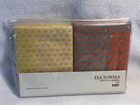 Two new HAY tea towels in original clear packaging. One towel has yellow dots on beige, other has orange dots on grey. Package label visible with product info.