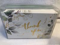 Front view of pack showing gold scripted "thank you" lettering on white card with green leafy botanical illustrations and gold foil accents.