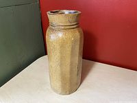 Full view of tall yellow ware stoneware jar with panelled sides, standing on a wood table against a red wall and green cabinet.