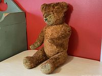 Medium brown plush bear posed sitting on a surface in front of a red wall, showing velvet paws, velvet snout, and glass eyes, straw-stuffed body.