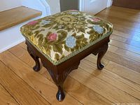 Angled view of foot stool showing the embroidered cushion top and carved wood legs on hardwood floor.