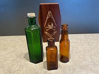 Overview of three antique poison bottles and a wooden mini coffin box with Mason symbols on a beige surface against a gray background.