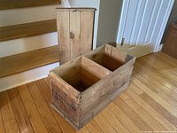 Antique wooden storage box with lid standing upright behind the box, placed on hardwood floor near staircase.